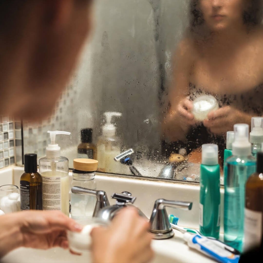 Menopause Skin Changes and How to Care for It Cluttered steamy bathroom sink: hands smear creams on face in smudged mirror reflection, battling dry skin.