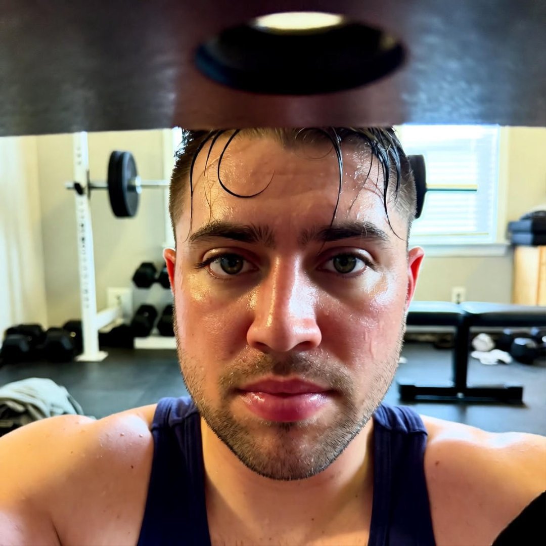 How to Lose Weight Fast Without Losing Muscle Mass? A sweaty, determined young man in a blue tank top reflects in a cluttered home gym mirror from a low crouch under a weight bench, capturing mid-workout resolve mixed with uncertainty.